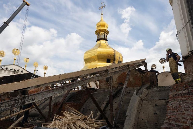 Workers in helmets stand amid rubble and wooden debris below a golden-domed church topped with a cross, with a crane visible against a partly cloudy sky.