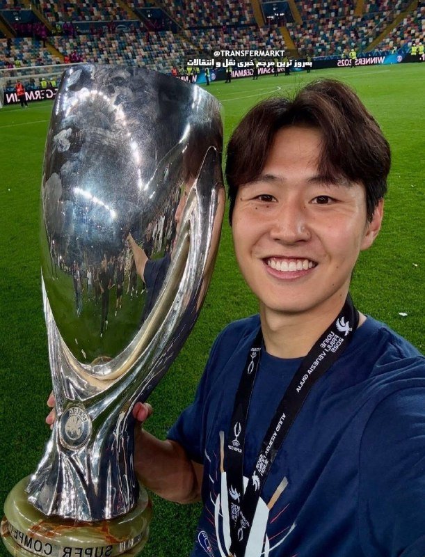 A smiling person takes a selfie holding a large silver trophy on a football pitch inside a stadium with "WINNERS 2023" and "@TRANSFERMARKT" visible on the screens behind.