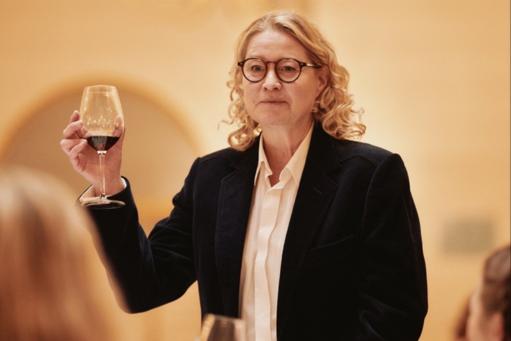 A woman with curly blonde hair and glasses, wearing a dark blazer over a white shirt, holds up a glass of red wine indoors.