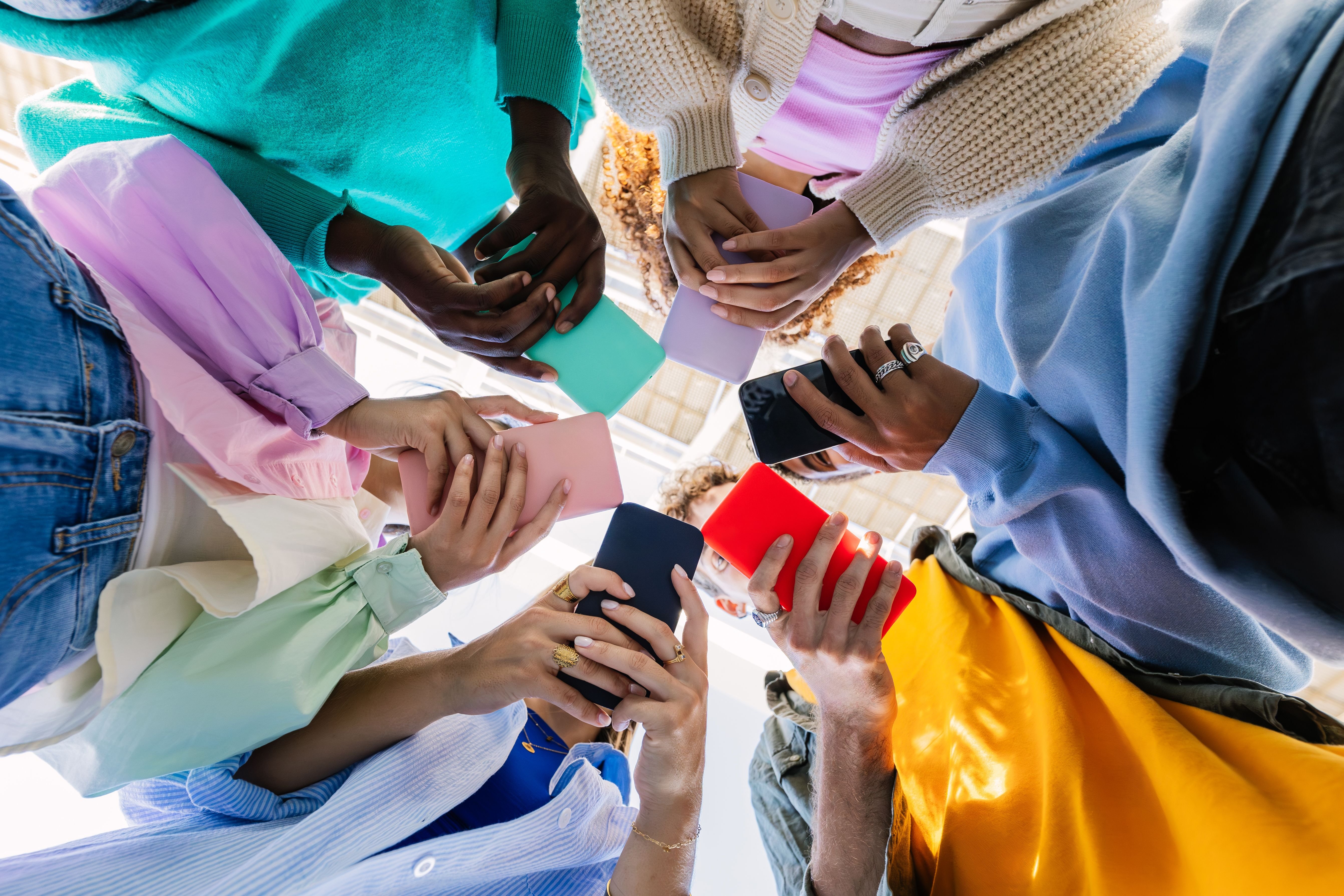 A diverse group of seven people stand in a circle viewed from below, each holding up colorful smartphones.