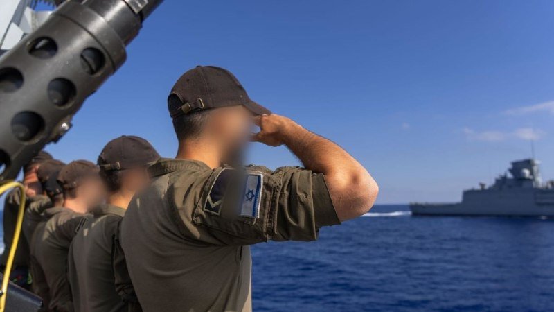Soldiers in olive uniforms with Israeli flag patches salute from a vessel as a naval ship sails on the open sea under a clear blue sky.