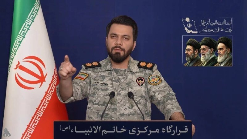 A bearded man in a camouflage military uniform gestures while speaking at a podium, with an Iranian flag on the left and a framed graphic of three bearded figures on the right.