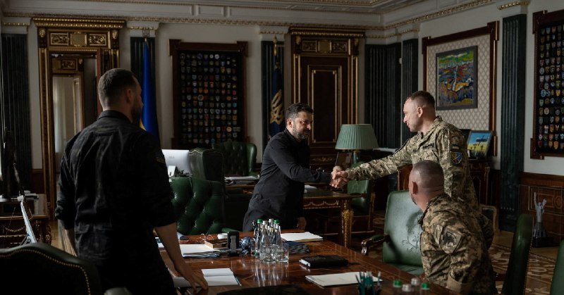 A man in a black shirt shakes hands with a man in military camouflage in an ornate, dark-wood office paneled with insignia displays.