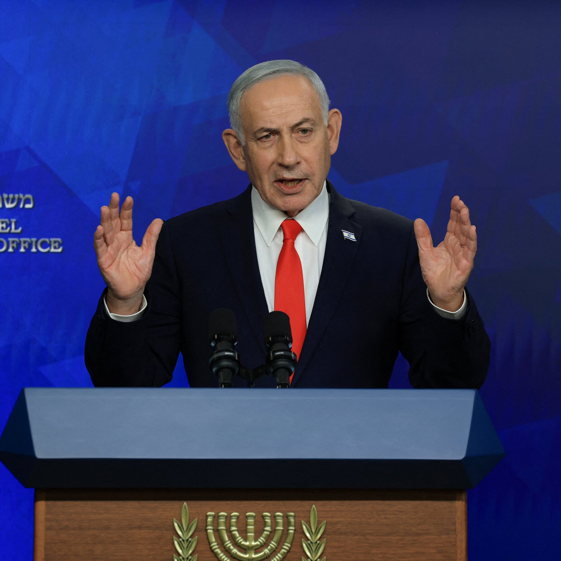 A gray-haired man in a dark suit and red tie gestures with both raised hands while speaking at a podium against a blue backdrop displaying a menorah emblem and Hebrew text.