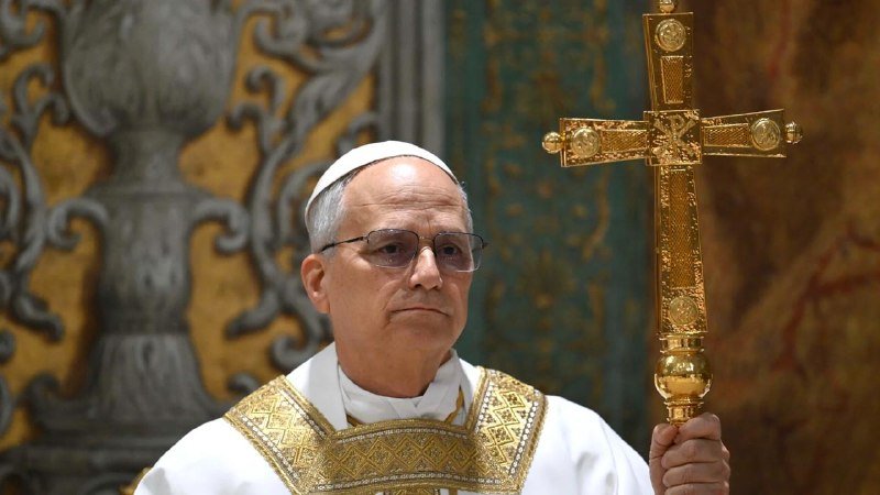 A Catholic clergyman in white and gold vestments and a zucchetto holds an ornate gold cross during a religious ceremony.