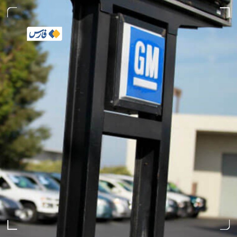 A black signpost displays the blue and white "GM" logo in the foreground, with parked cars and a building visible in the blurred background.