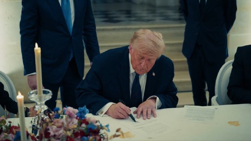 A man in a dark suit signs a document at a candlelit table while others stand behind him in a formal setting.