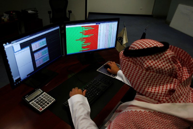A person in traditional Arab headdress works at a desk with dual monitors displaying red and green financial data, alongside a keyboard and calculator.
