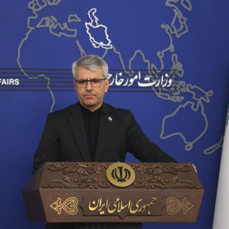 A gray-haired man in a dark suit and glasses stands at a wooden podium bearing an Iranian emblem, with a blue world map backdrop displaying Persian script.