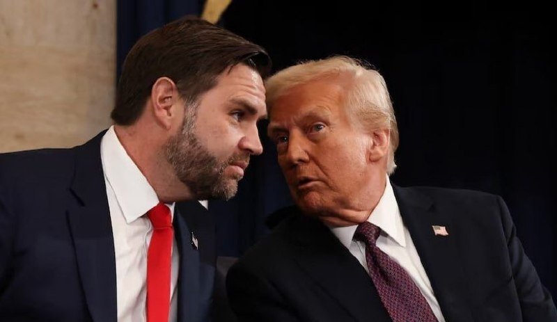 Two men in dark suits sit side by side, leaning close as if conversing, against a dark backdrop with partial gold trim visible behind them.