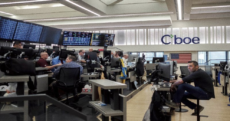 Traders work at computer terminals beneath a large "Cboe" sign and digital display boards in a busy trading floor, with an American flag visible in the background.