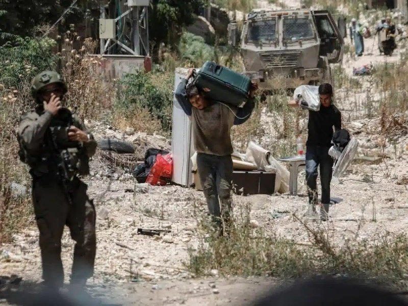 Two men carry suitcases and bags through a debris-strewn outdoor area while an armed soldier in uniform stands on the left, with an armored military vehicle visible in the background.
