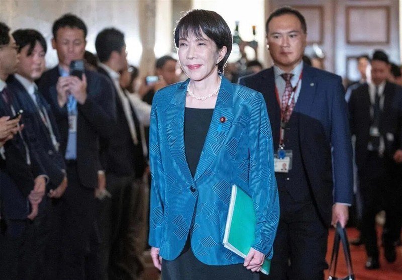 A woman in a blue blazer with a pearl necklace and lapel pin walks through a hallway, holding a green folder, surrounded by men in suits and onlookers.