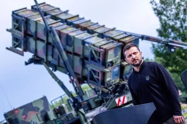 A bearded man in a dark shirt stands at a podium with microphones in front of a camouflaged surface-to-air missile system.