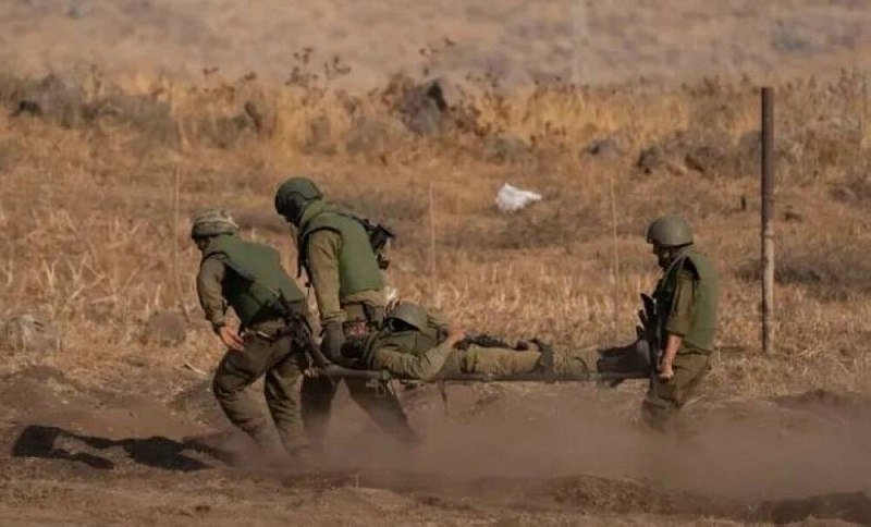 Four soldiers in helmets and green uniforms carry a wounded comrade on a stretcher across a dusty, dry field.