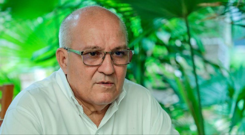 A balding older man wearing glasses and a white shirt sits in front of lush green foliage, looking slightly off-camera.