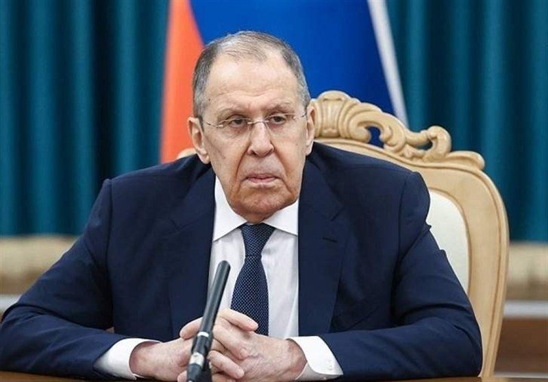 A man in a dark suit and tie sits at a table with a microphone, positioned before a tricolor flag and an ornate chair.