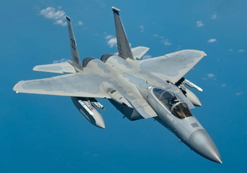 Two gray fighter jets fly in close formation against a blue sky, their cockpits visible and external fuel tanks mounted beneath the wings.