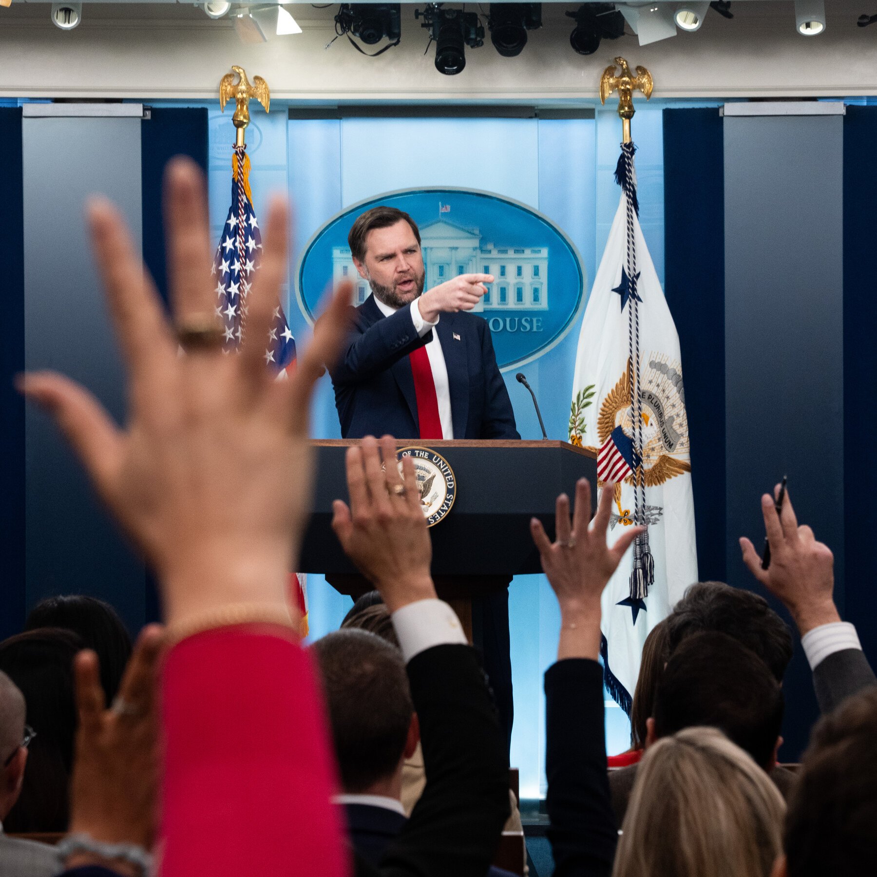 A bearded man in a dark suit and red tie stands at a podium bearing the presidential seal, pointing toward reporters with raised hands during a White House press briefing.