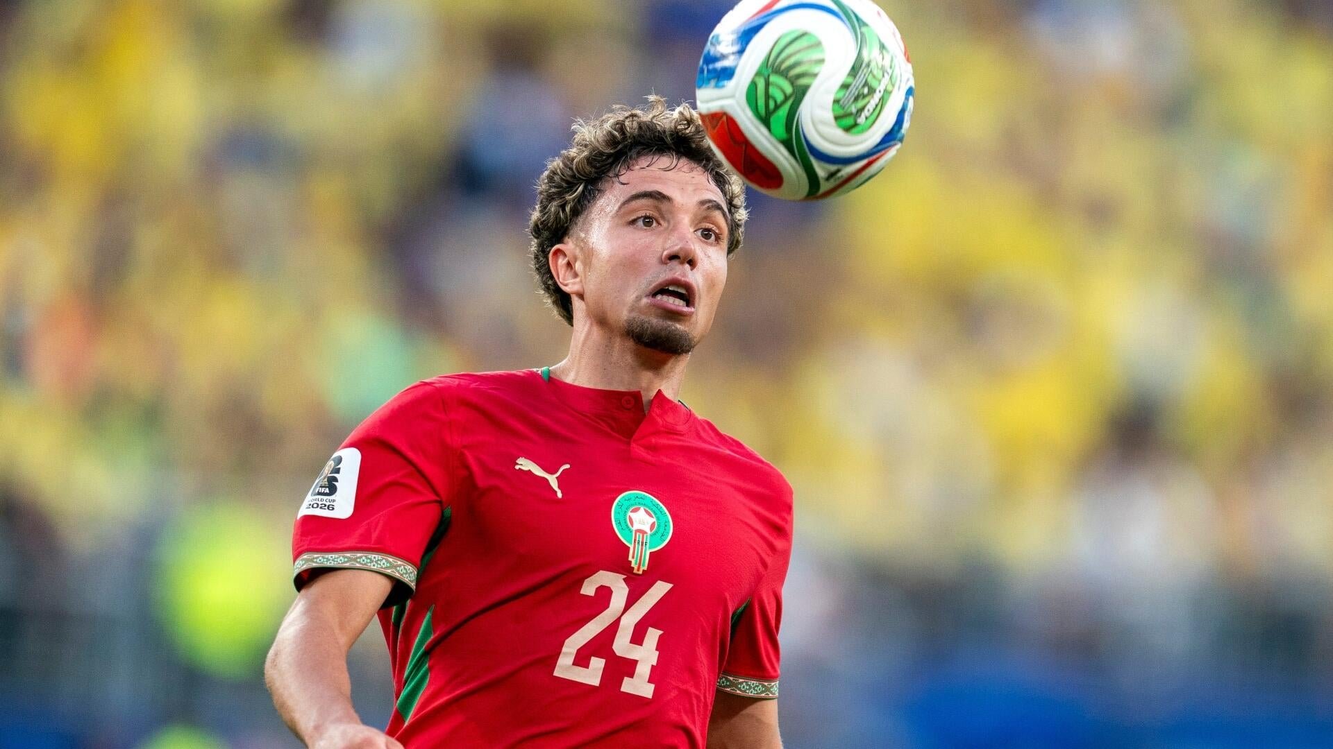 A soccer player in a red Morocco jersey with the number 24 heads a colorful ball during a match in a stadium filled with yellow-clad spectators.