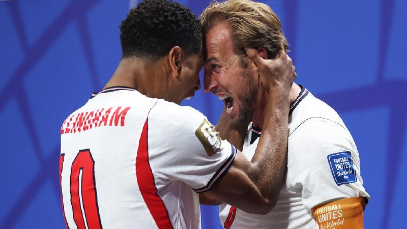 Two soccer players in white jerseys with red and gold accents embrace head-to-head, one shouting joyously while gripping the other's face, against a blue background.