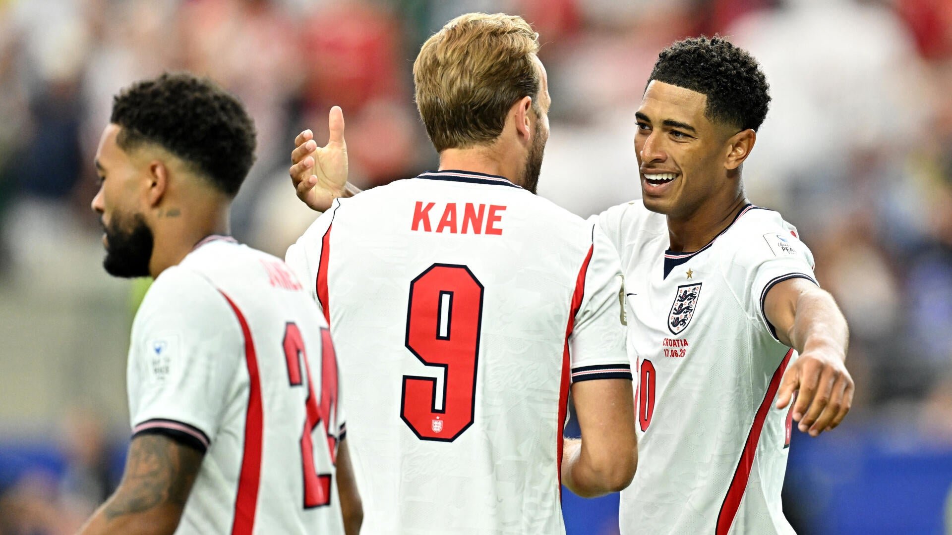 Three England soccer players in white jerseys celebrate, with one wearing a jersey labeled "KANE" and the number 9, set against a blurred stadium crowd.