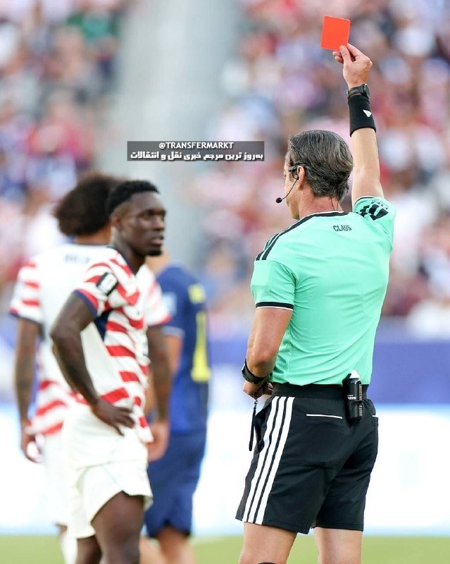 A referee in a green shirt holds up a red card toward players wearing red-and-white striped jerseys during a soccer match in a stadium.