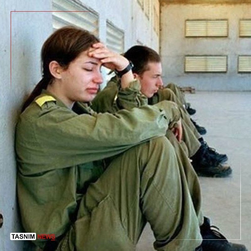 Women in green military uniforms sit against a building wall, with one resting her head in her hand in an outdoor courtyard.