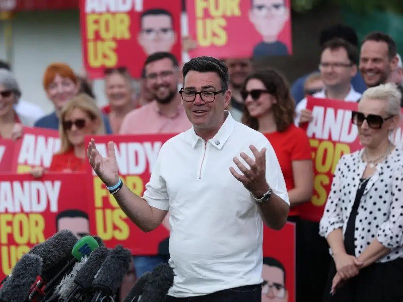A man in a white shirt and glasses stands before microphones, speaking with raised hands while supporters behind him hold red "Andy For Us" campaign signs featuring illustrated portraits.