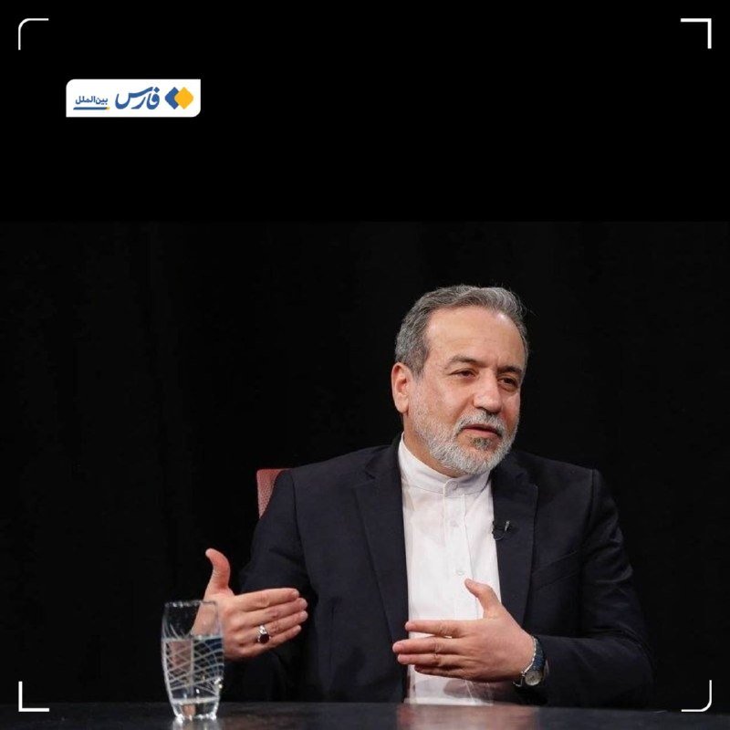 A gray-bearded man in a dark suit and white shirt gestures with his hands while seated at a table with a glass of water, against a black background with Persian text in the corner.