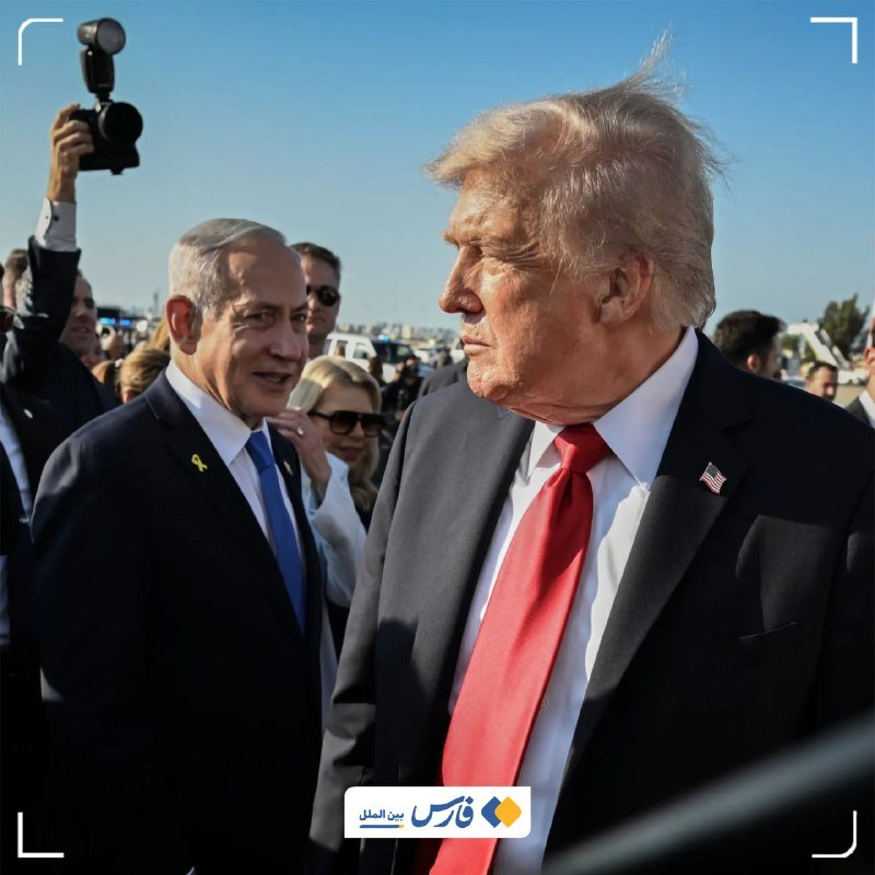 Two men in dark suits stand outdoors in a crowd, one wearing a red tie and a US flag pin, the other a blue tie with a yellow ribbon, as a photographer holds up a camera nearby.