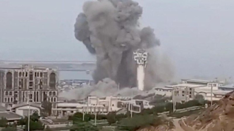 A large plume of gray smoke rises from an explosion near a waterfront industrial area, with damaged buildings and a communications tower visible in the foreground.