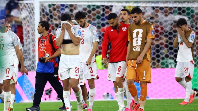 Soccer players in white and red uniforms walk dejectedly off the pitch past a goal net, accompanied by a goalkeeper in orange and a staff member with a camera.
