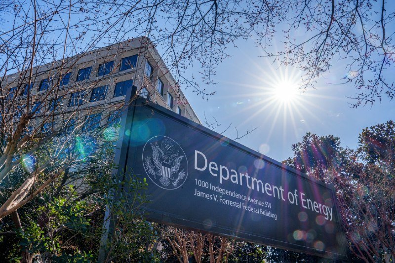 A sign reading "Department of Energy, 1000 Independence Avenue SW, James V. Forrestal Federal Building" stands before a glass office building under a bright sunlit sky.