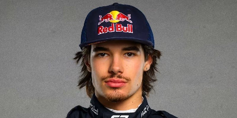 Young man wearing a navy Red Bull racing cap and matching team apparel poses for a portrait against a gray background.