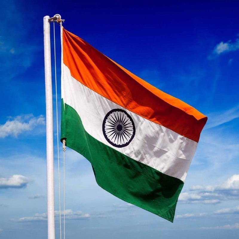 The Indian national flag, featuring saffron, white, and green horizontal stripes with a navy blue wheel in the center, flies from a white flagpole against a blue sky with scattered clouds.
