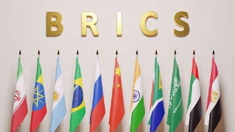 Ten national flags stand in a row beneath the gold letters "BRICS" mounted on a light-colored wall.