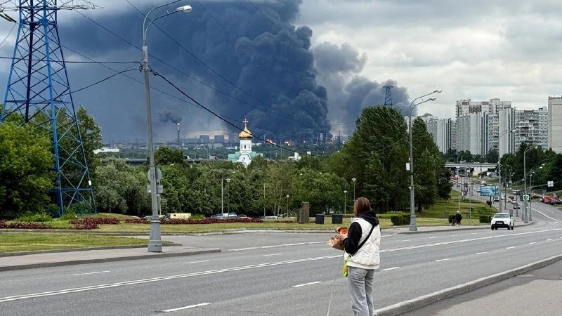 A person stands on a roadside holding a bag while thick black smoke billows from industrial structures in the distance, with high-rise apartment buildings visible to the right.
