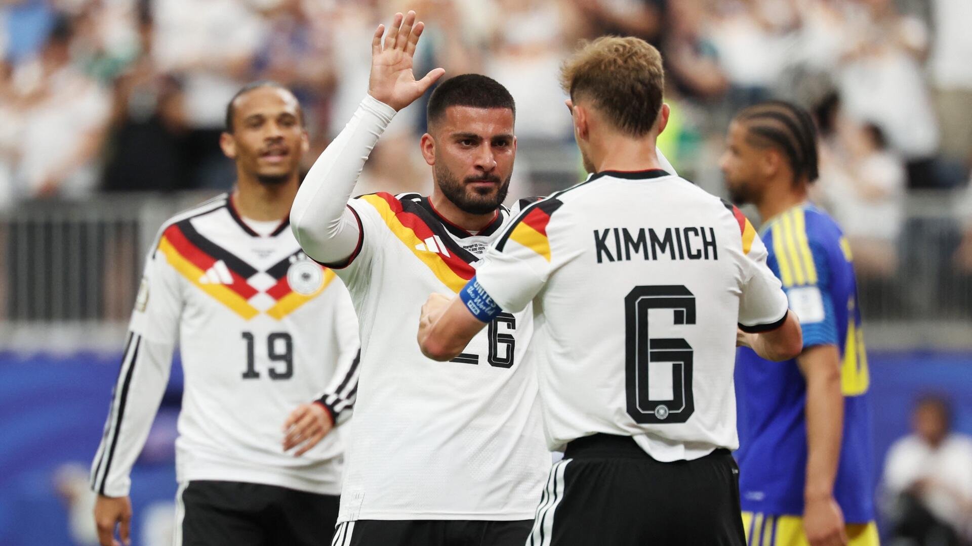 Two soccer players in white Germany jerseys, including number 6 "Kimmich," celebrate on the field during a match, with other players and a blurred crowd visible in the background.