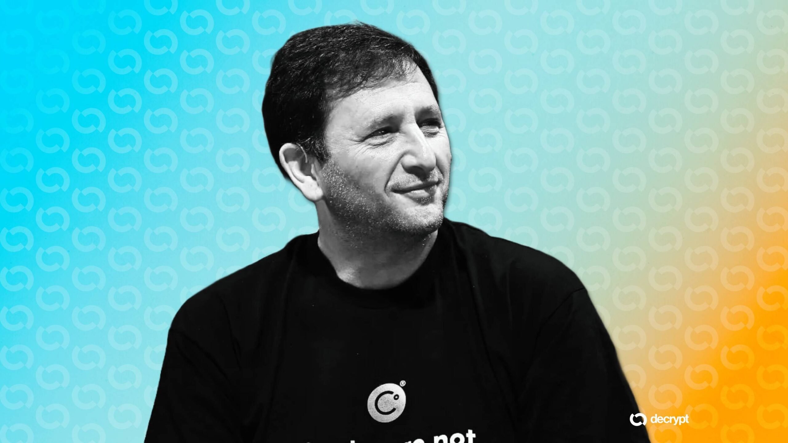 A man with short dark hair and stubble, wearing a black t-shirt, smiles while looking to the side against a teal and orange patterned background featuring a "decrypt" logo.
