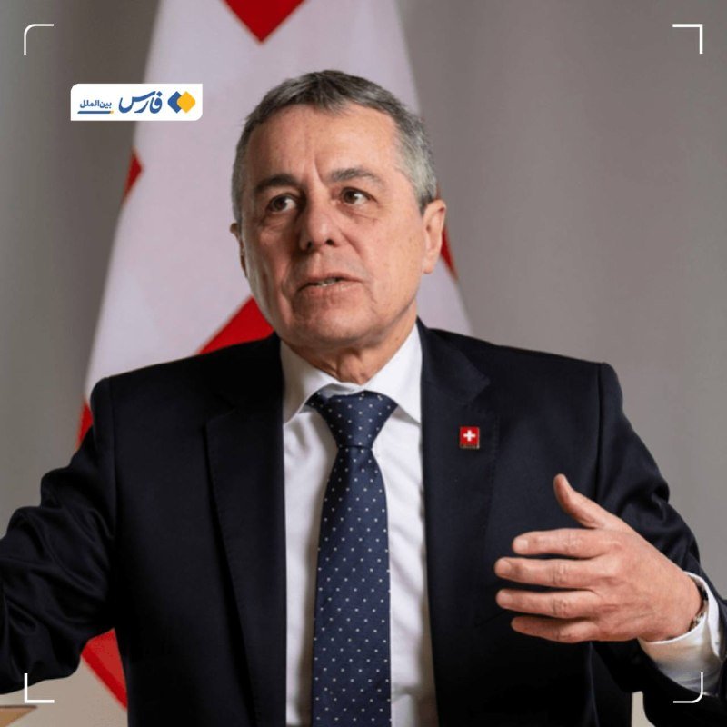 A gray-haired man in a dark suit and dotted tie speaks while gesturing with his hand, standing before a Swiss flag with a news channel logo visible in the corner.
