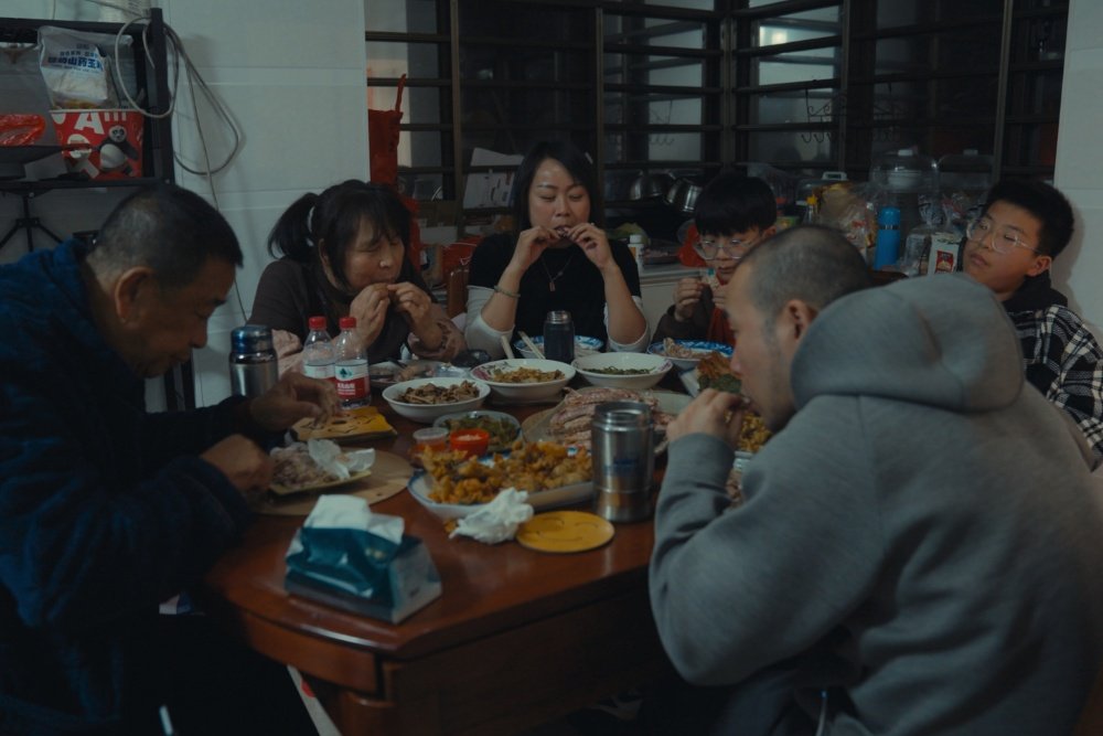 Six people gather around a table in a kitchen, eating a meal of various dishes including crab and a bowl of mixed food.