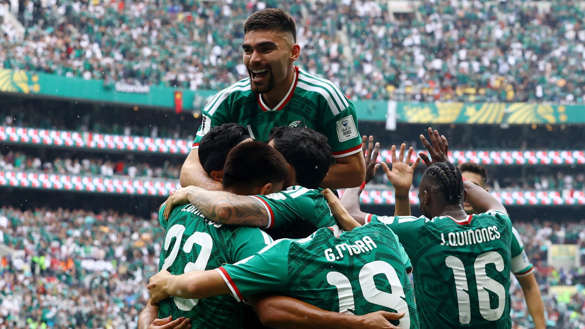 Soccer players in green Mexico jerseys, including numbers 22, 26, and 16 (J. Quiñones), embrace in a celebratory hug before a packed stadium crowd.