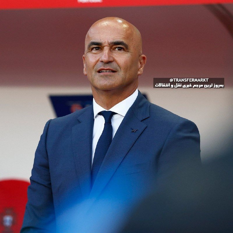 A bald man in a navy blue suit and tie stands on a red stadium sideline, looking forward.