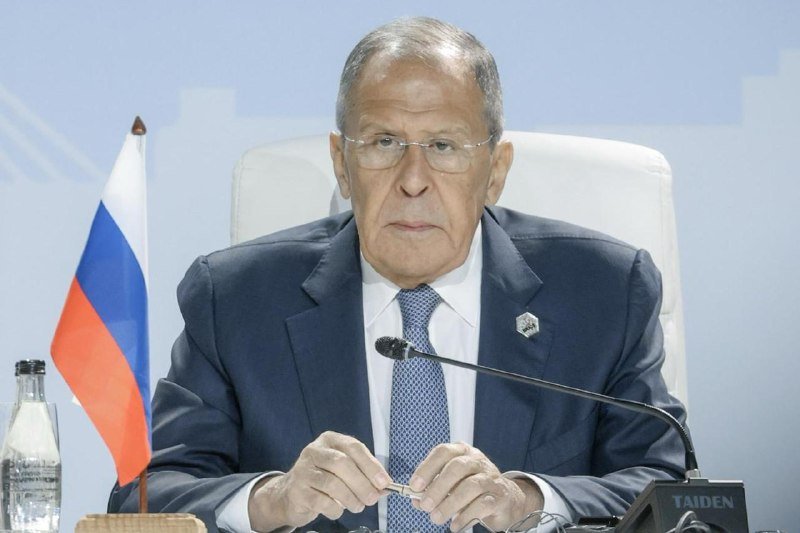 An older man in a blue suit and glasses sits at a conference table with a microphone, a Russian flag, and a TAIDEN branded device visible.