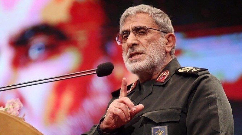 A gray-bearded man in a green military uniform with insignia speaks at a podium, gesturing with one finger raised in front of a microphone.