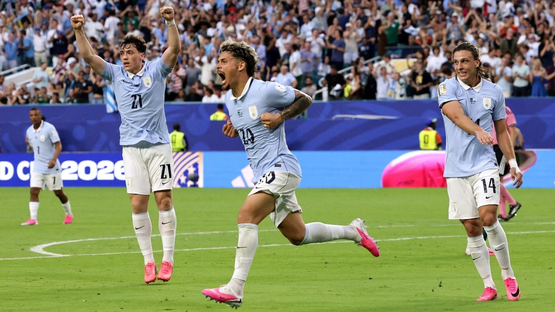 Three soccer players in light blue jerseys celebrate on the pitch, one jumping with arms raised and another shouting, while a large crowd cheers in the stadium behind them.