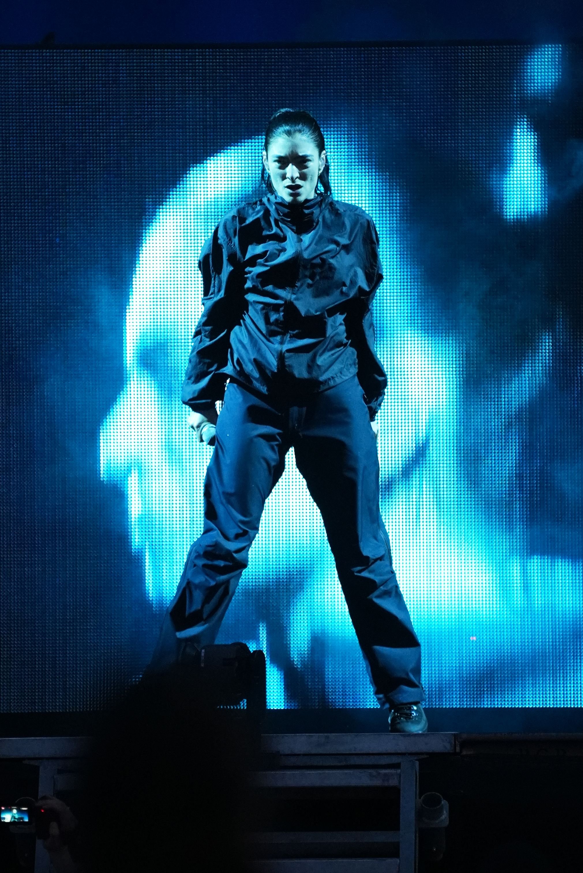 A performer in a dark jumpsuit stands on stage, holding a microphone, illuminated by blue stage lighting against a large LED screen backdrop.