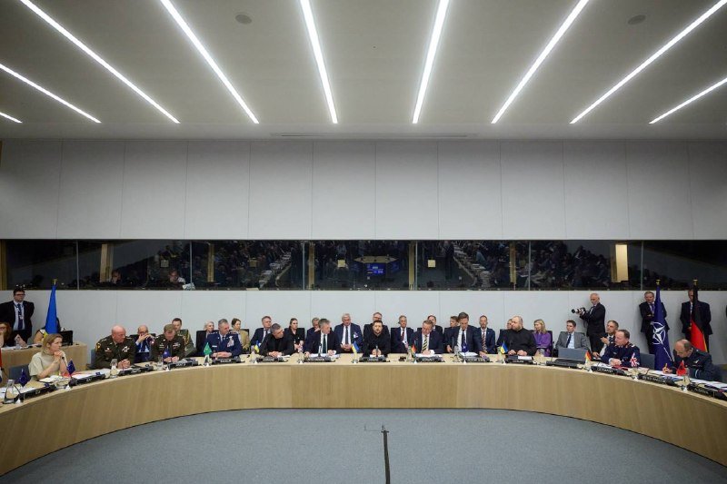 Officials in suits and military uniforms are seated around a large circular conference table beneath bright linear ceiling lights, with flags visible and observers behind a glass panel.