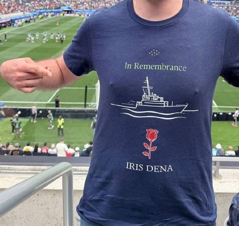 A person wearing a navy shirt reading "In Remembrance" with a ship graphic, a red rose, and "IRIS DENA" stands in a stadium overlooking a soccer match.